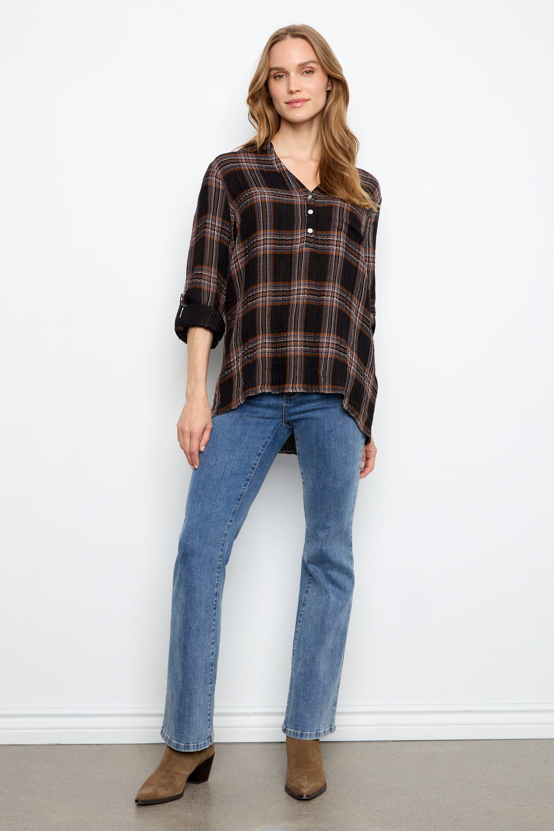A woman modeling a dark brown and black plaid button-down shirt with three-quarter sleeves, paired with blue jeans and brown ankle boots, standing against a white wall.