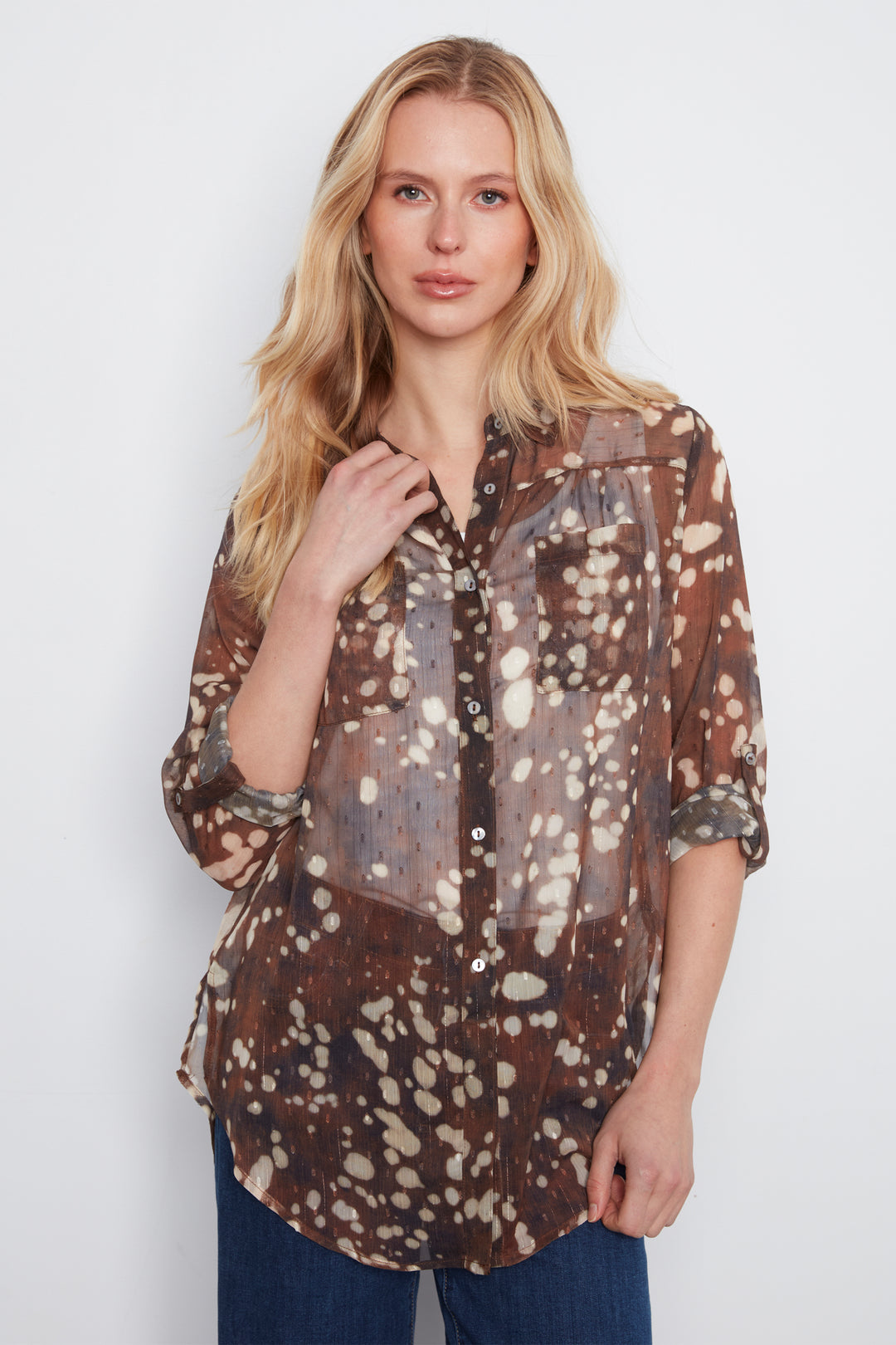 A woman modeling a sheer brown button-up blouse with a white and beige speckled tie-dye pattern, paired with dark blue jeans, against a plain light gray background.
