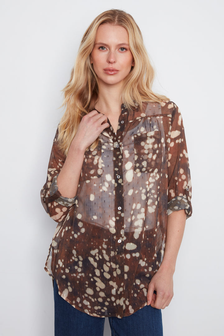 A woman modeling a sheer brown button-up blouse with a white and beige speckled tie-dye pattern, paired with dark blue jeans, against a plain light gray background.