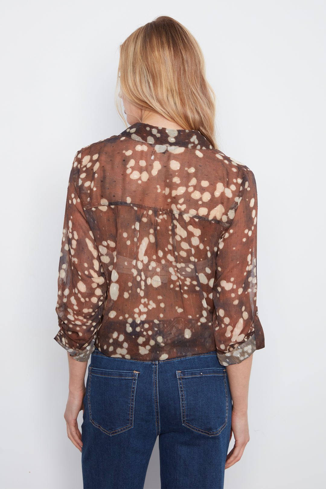 A woman viewed from the back wearing a sheer brown long-sleeve blouse with a beige and white spotted pattern, paired with dark blue jeans, against a plain white background.