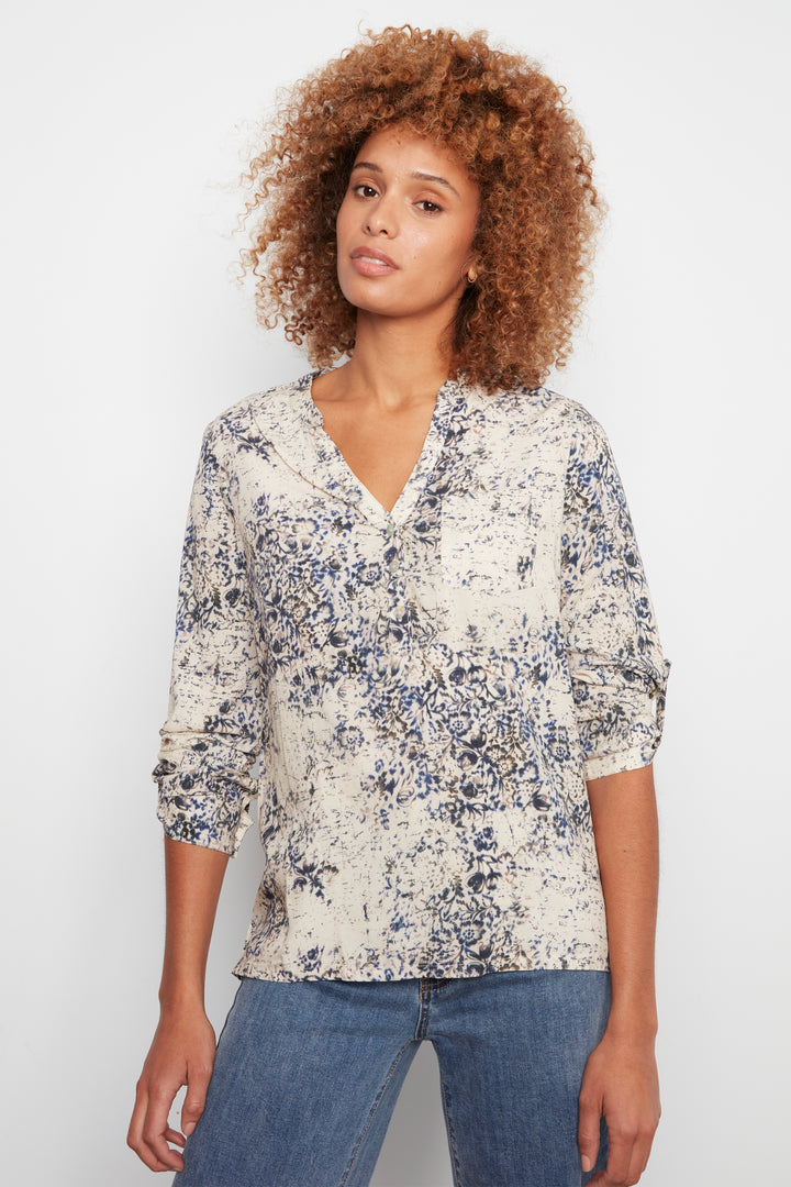 A woman with curly light brown hair wearing a cream-colored long-sleeve blouse with a blue and gray floral pattern, paired with blue jeans, against a plain white background.