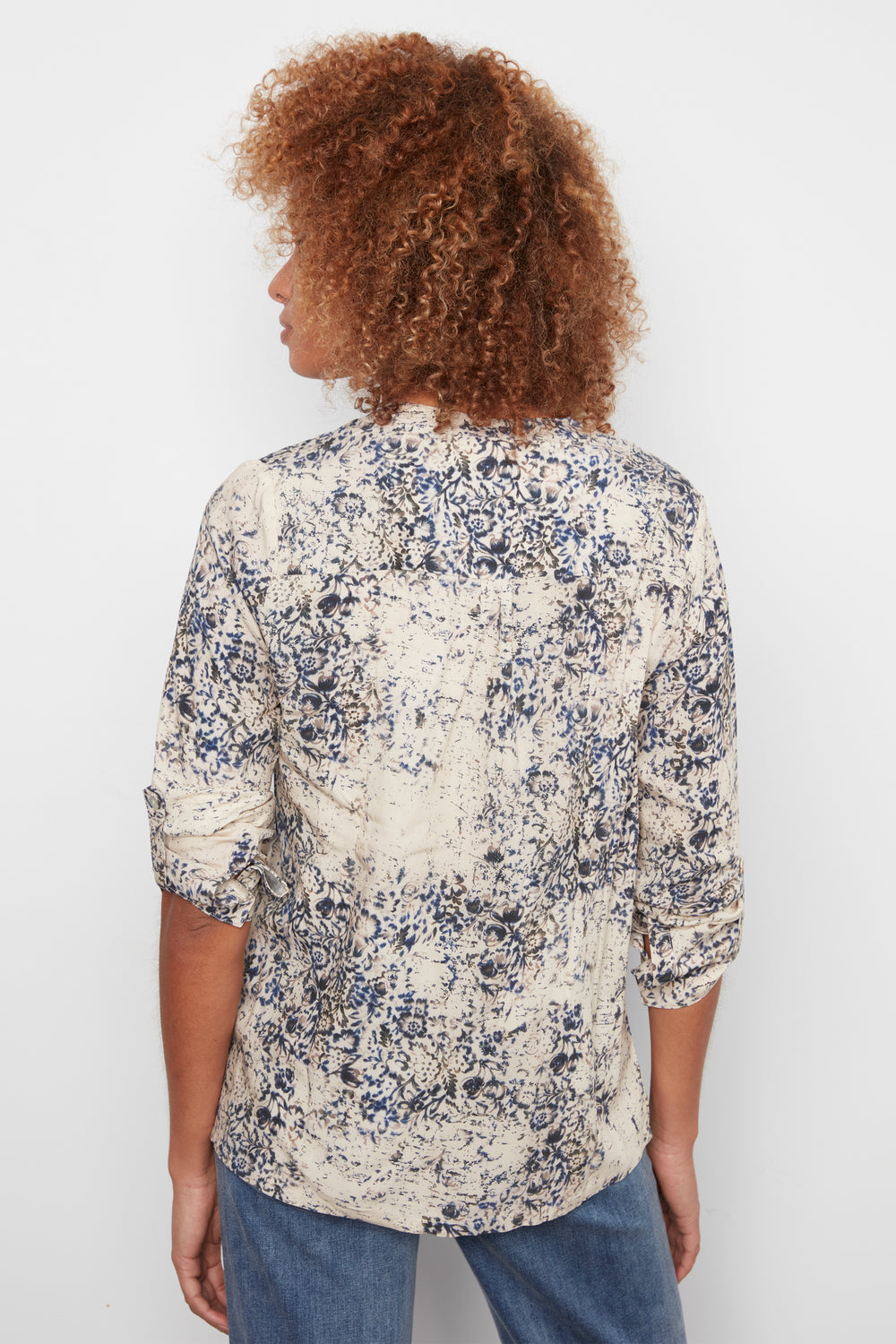 A woman with curly brown hair is shown from the back wearing a cream-colored button-up shirt with a blue and gray abstract floral pattern, paired with blue jeans, against a plain white background.