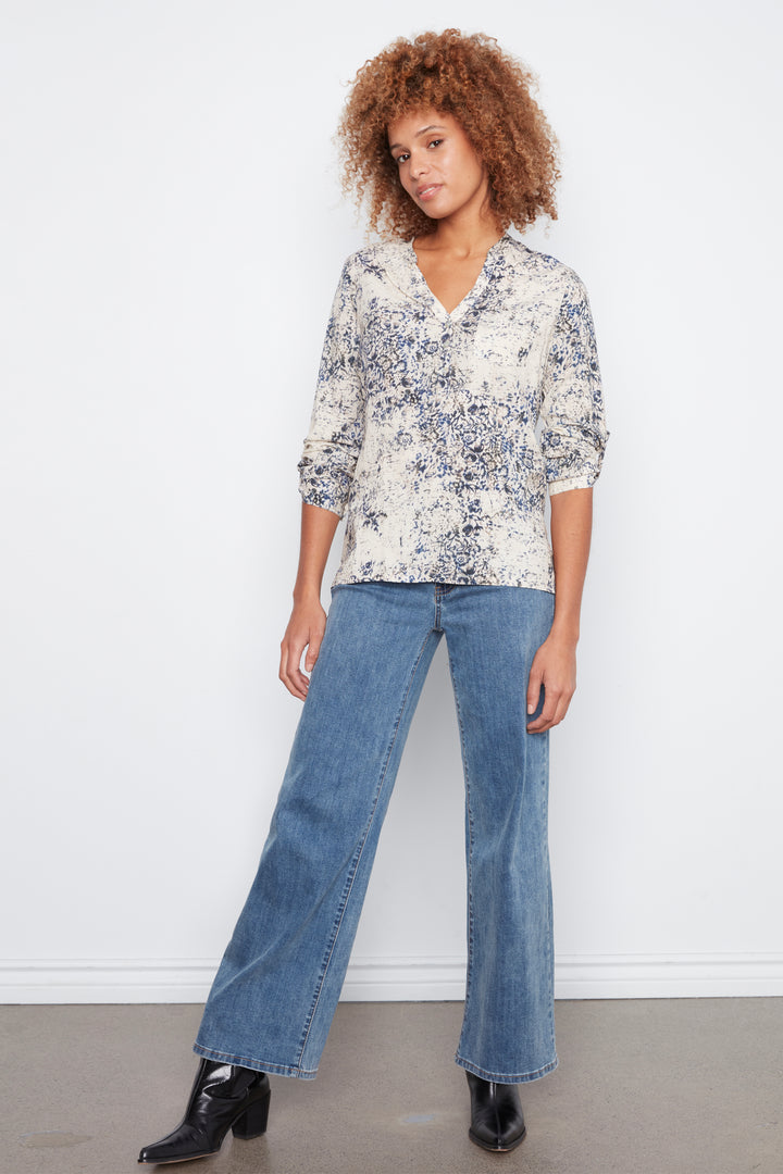 A woman modeling a three-quarter sleeve button-up blouse with a blue and white abstract floral pattern, paired with wide-leg blue jeans and black ankle boots, standing against a white wall.