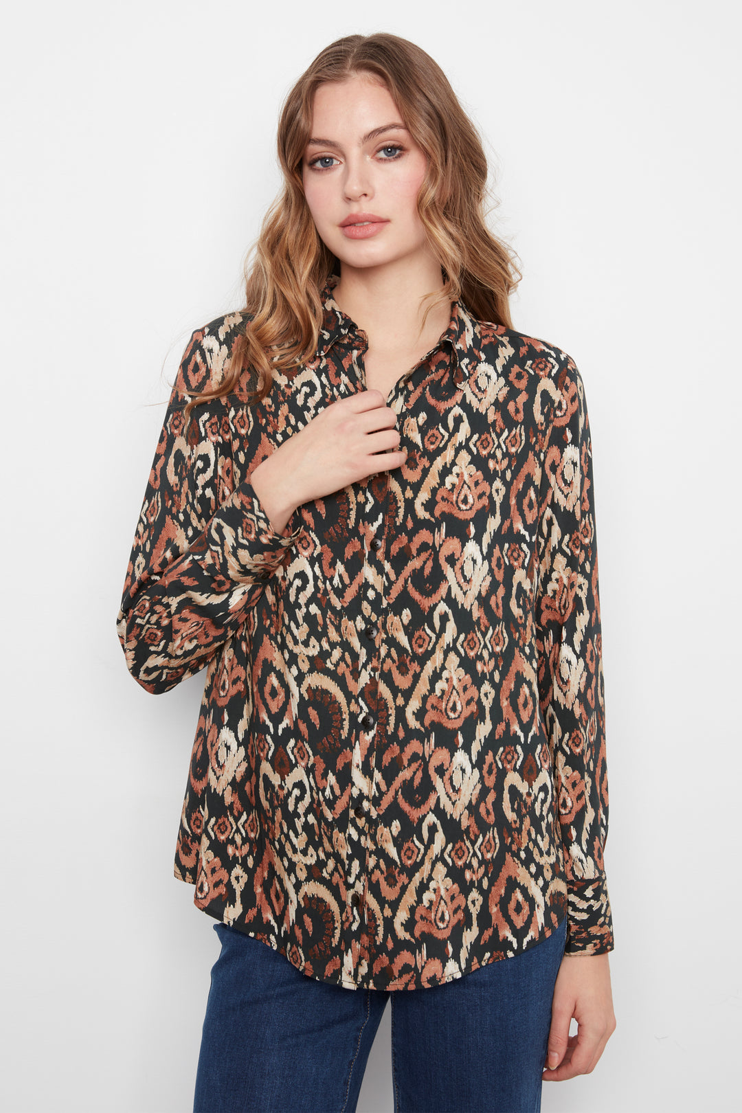 A woman wearing a long-sleeve button-up blouse with an intricate abstract pattern in black, rust orange, and beige, paired with dark blue jeans, photographed against a light gray studio background.