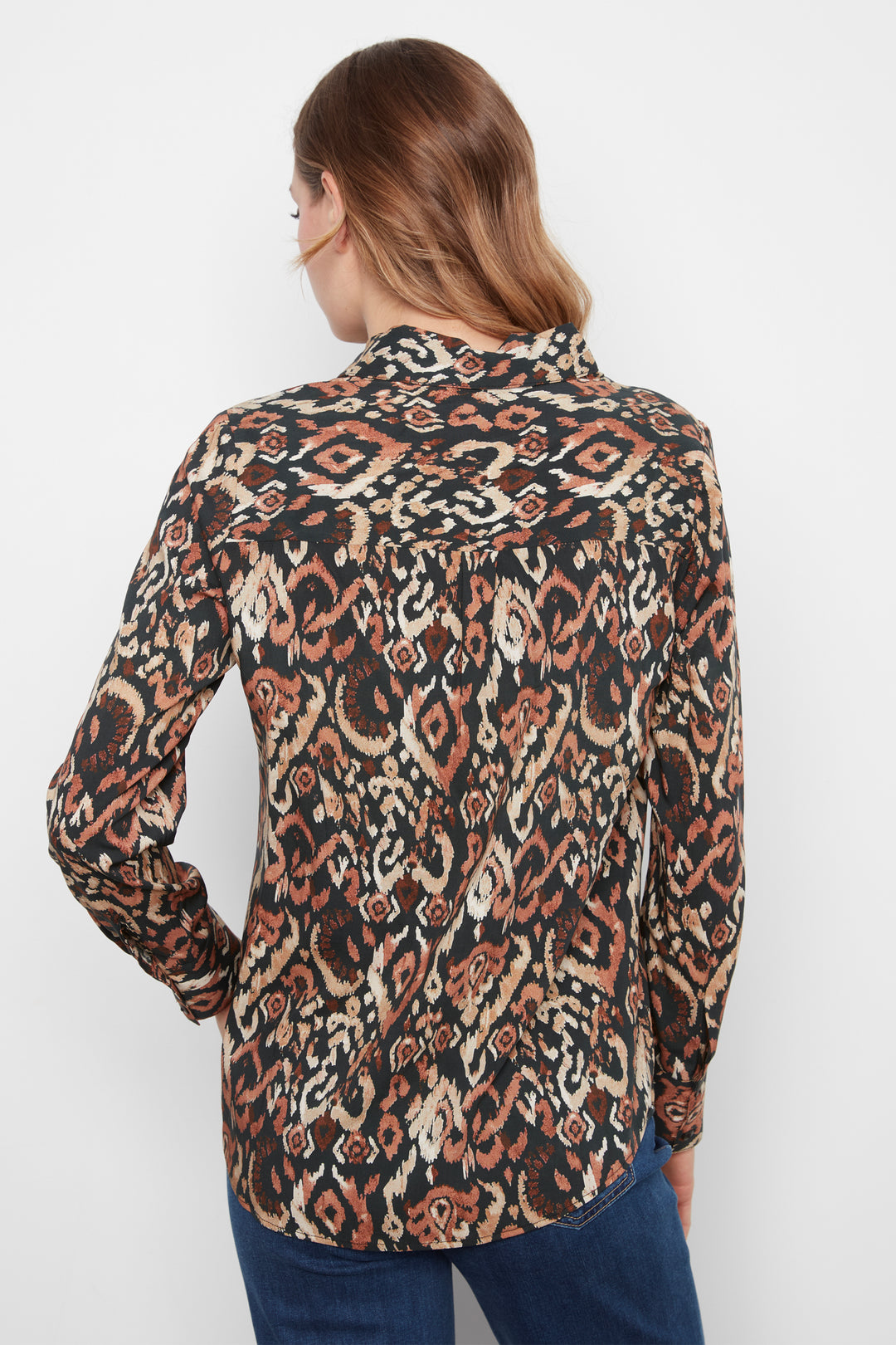 A woman viewed from the back wearing a long-sleeve button-up shirt with an intricate brown, beige, and black geometric pattern, paired with blue jeans against a plain white background.