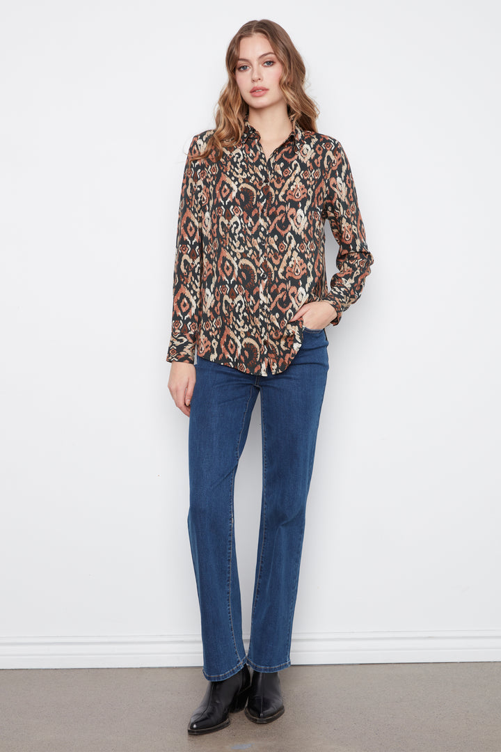 A woman modeling a long-sleeve button-up shirt with a brown, beige, and black geometric pattern, paired with blue jeans and black boots, standing against a white wall.