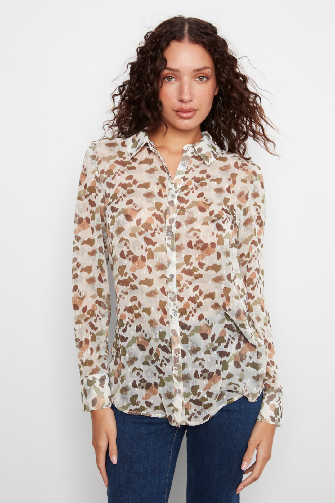 A woman wearing a long-sleeve button-up blouse with an abstract brown and tan pattern on a cream background, paired with dark blue jeans, against a plain white studio background.