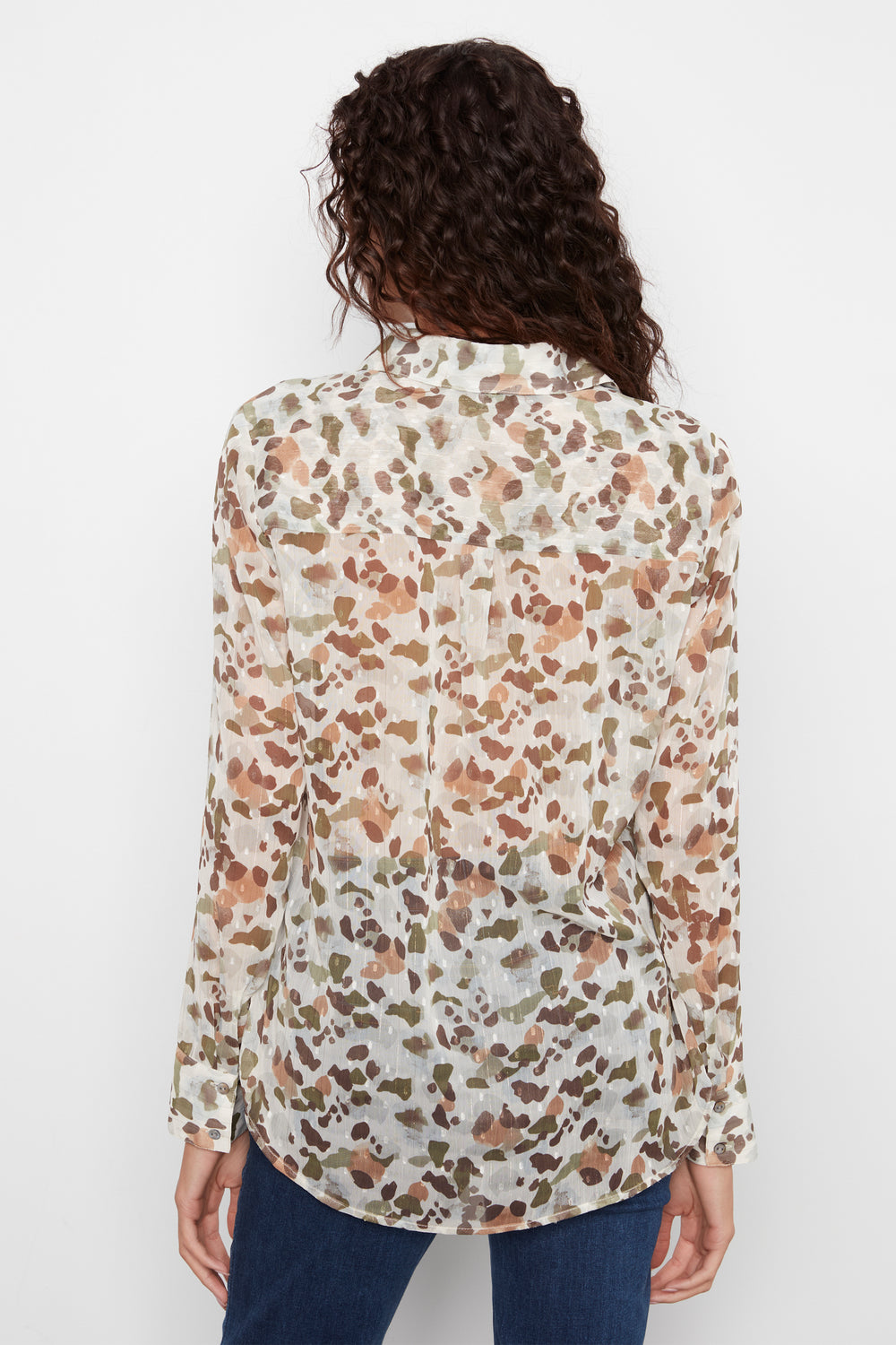 A woman viewed from the back wearing a long-sleeve button-up blouse with an abstract watercolor-style print in brown, beige, and muted green on an off-white base, paired with dark blue jeans against a plain white background.