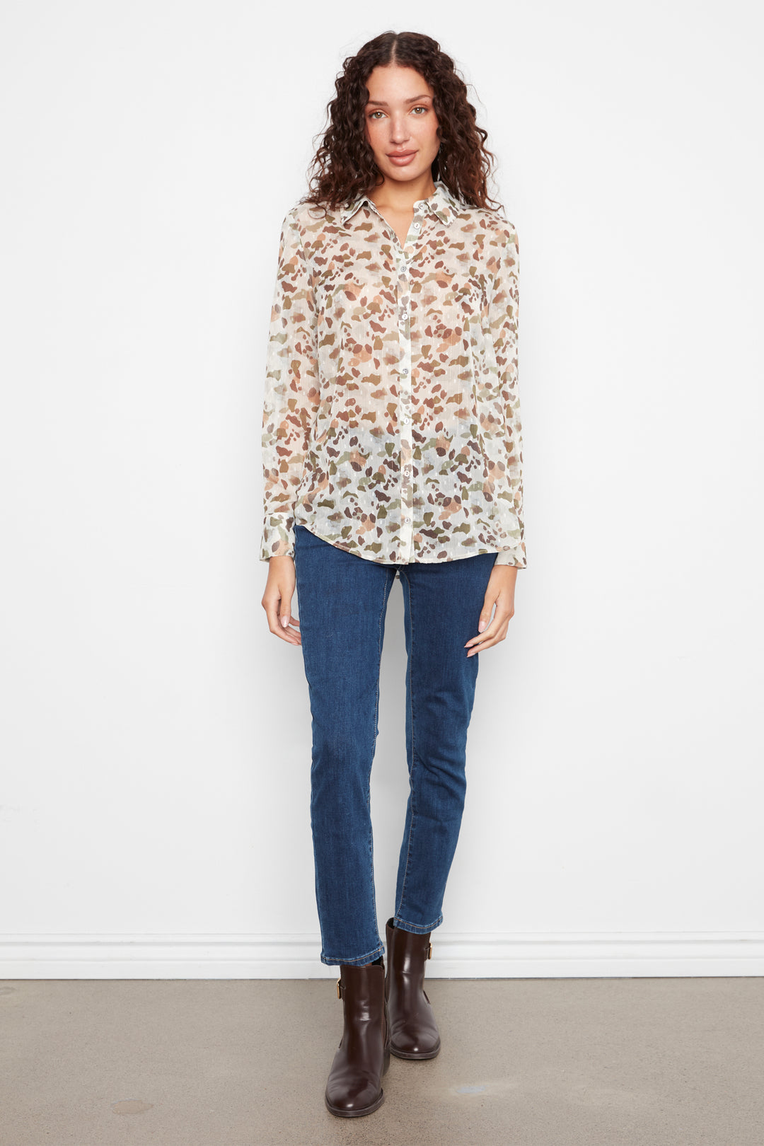 A woman stands against a plain white wall wearing a long-sleeve button-up blouse with a brown and beige floral print, dark blue jeans, and dark brown ankle boots.