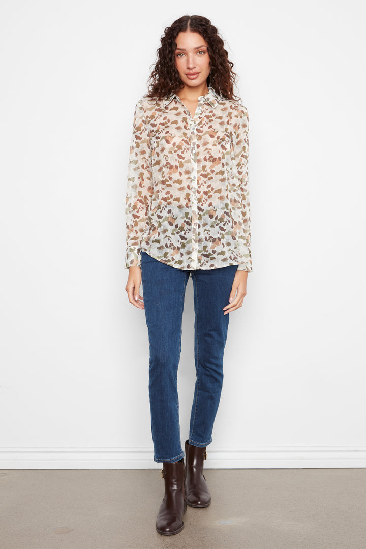 A woman stands against a plain white wall wearing a long-sleeve button-up blouse with a brown and beige floral print, dark blue jeans, and dark brown ankle boots.