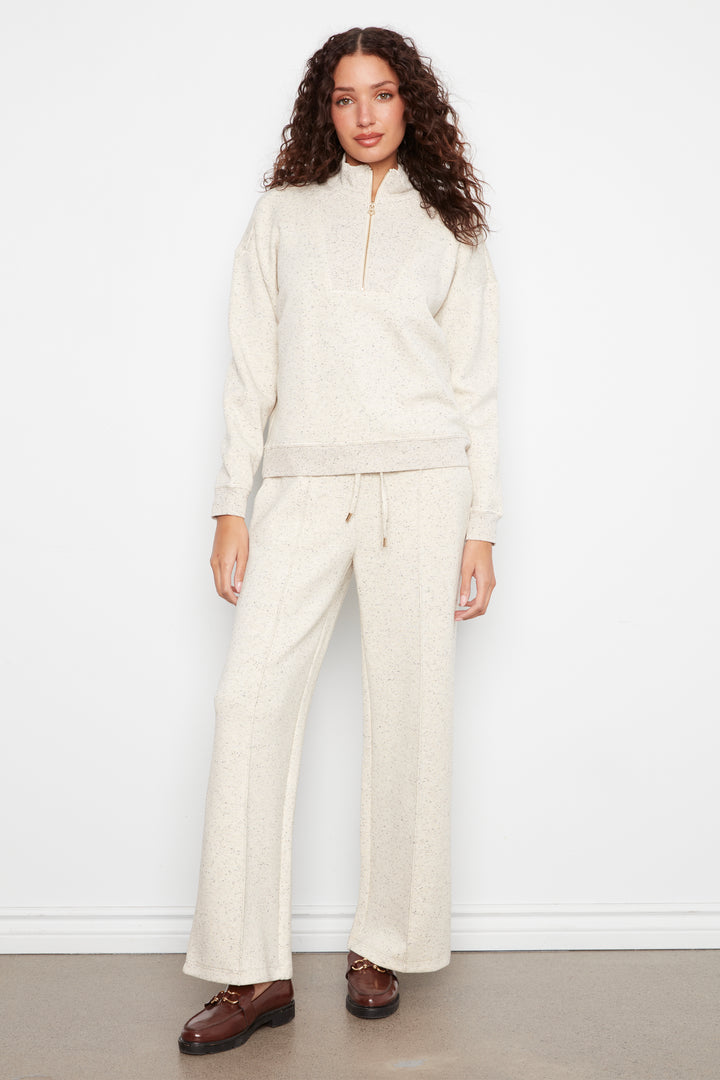 A woman modeling a matching cream-colored knit lounge set consisting of a half-zip pullover and wide-leg pants, paired with brown leather shoes, against a plain white wall.