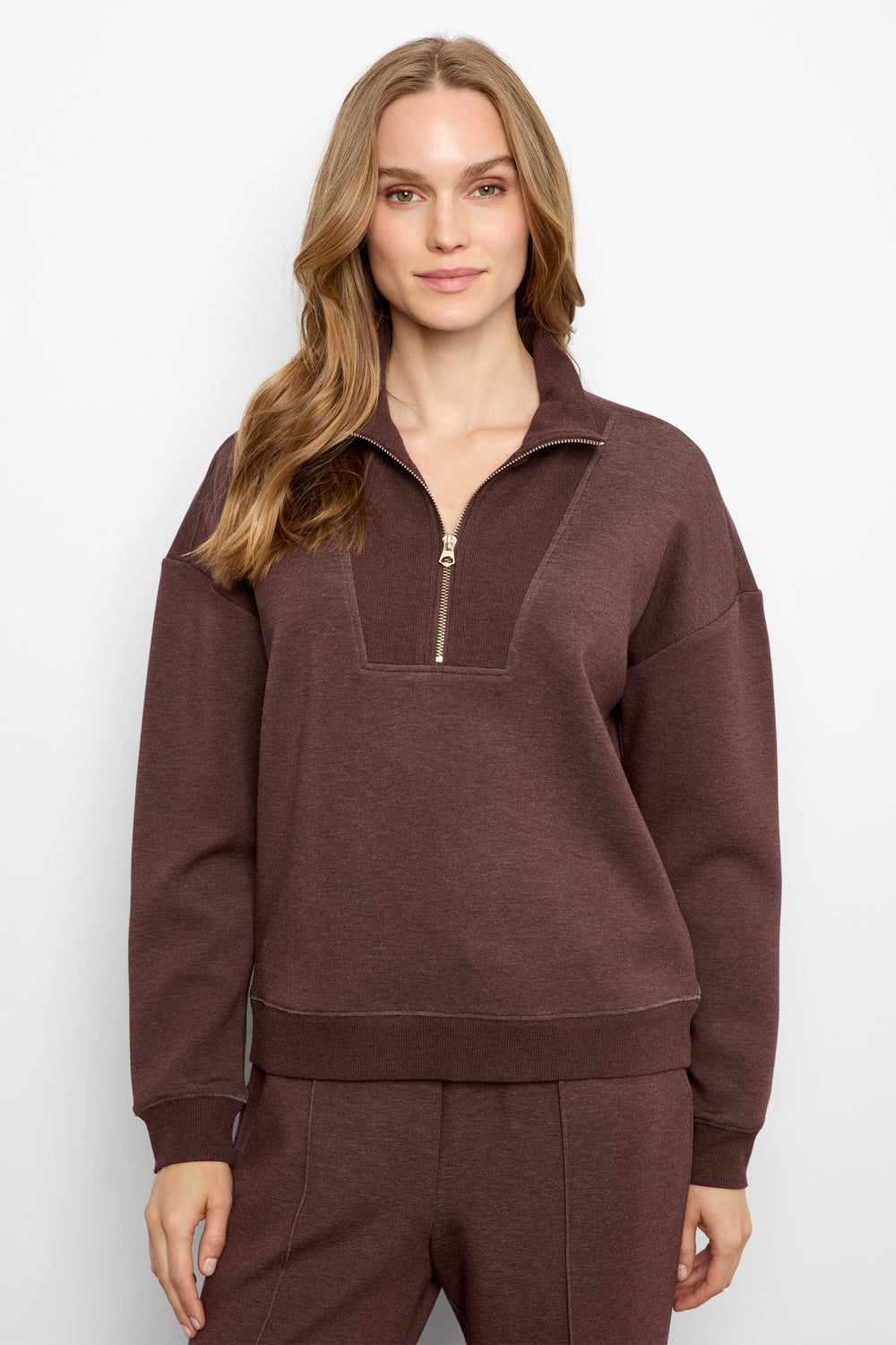 A woman modeling a dark brown half-zip sweatshirt with a matching pair of sweatpants against a light gray studio background.