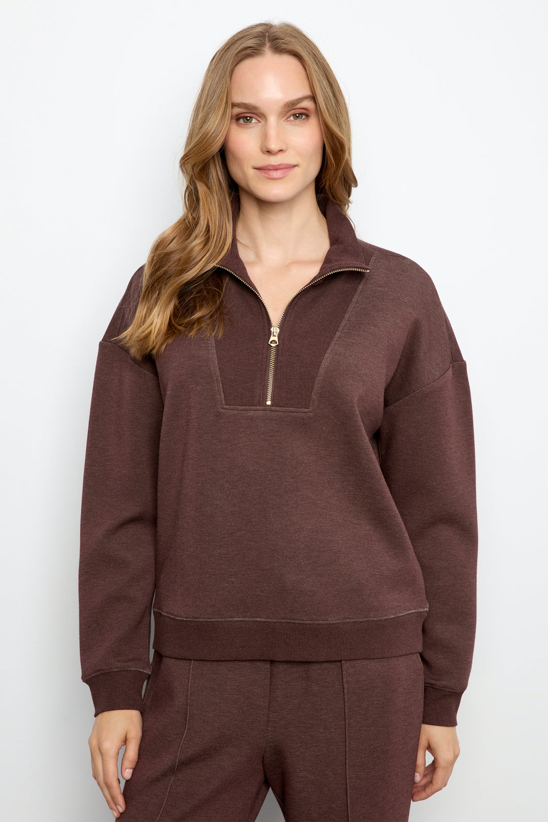 A woman modeling a dark brown half-zip sweatshirt with a matching pair of sweatpants against a light gray studio background.