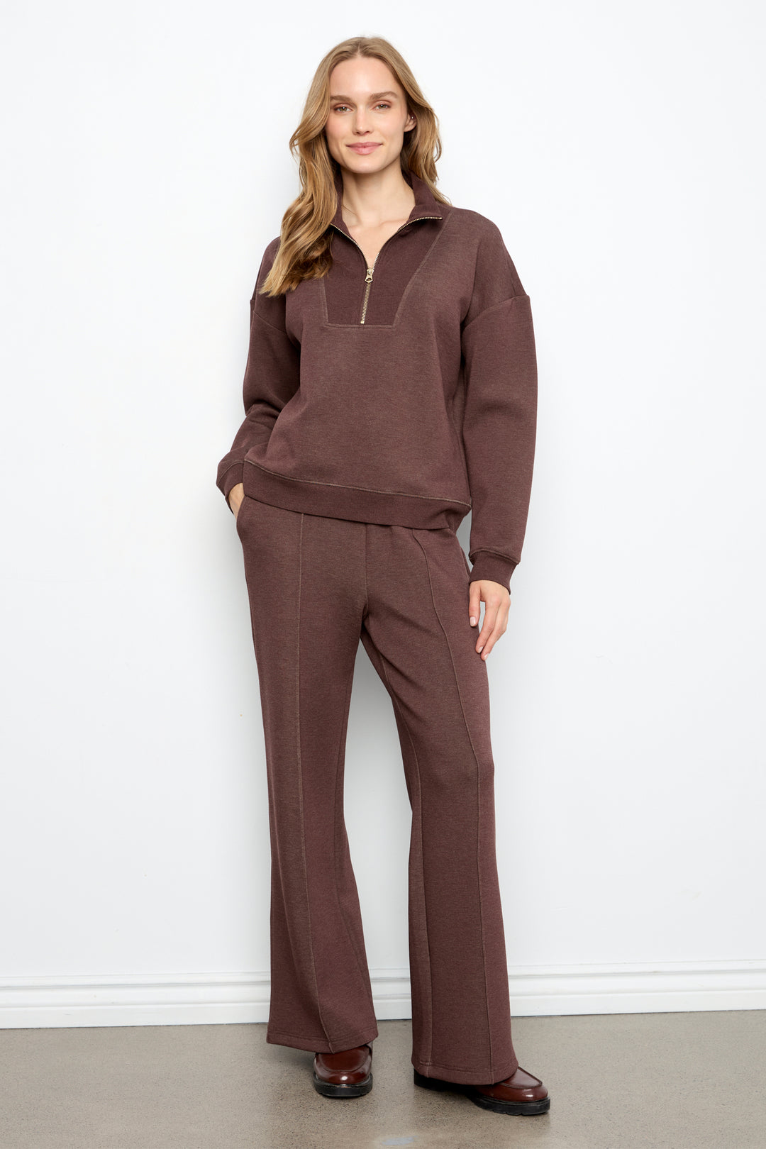 A woman modeling a matching brown lounge set consisting of a long-sleeve half-zip pullover and wide-leg pants, standing against a plain white wall.