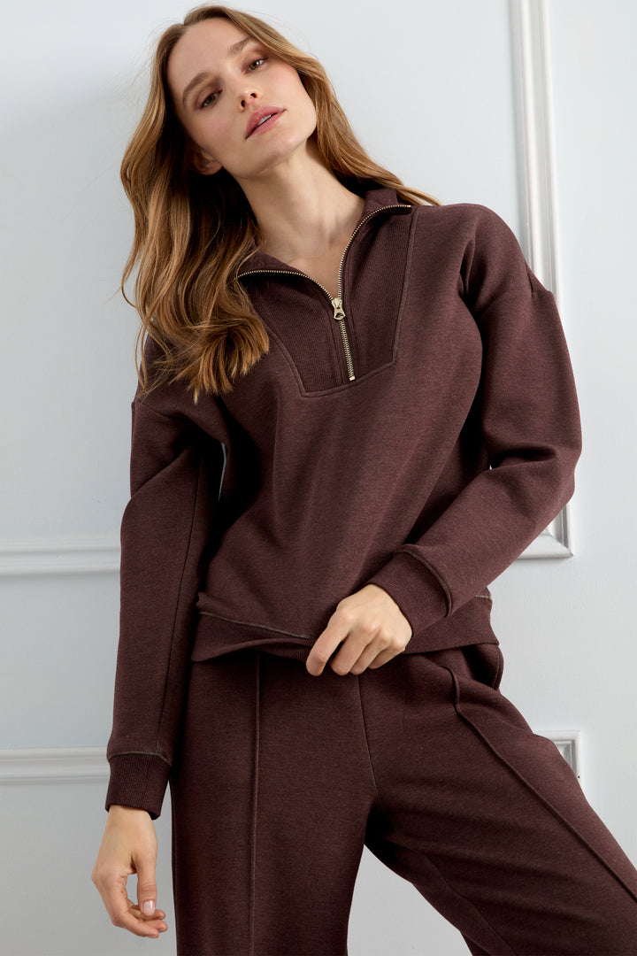 A woman modeling a matching dark brown lounge set consisting of a half-zip sweatshirt and coordinating sweatpants, set against a light gray paneled wall.
