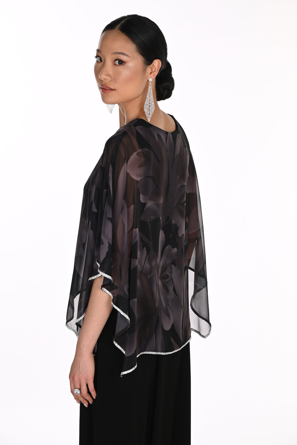 A woman shown from the side and back wearing a sheer black floral chiffon cape top with white trim over a black dress, against a plain white background.