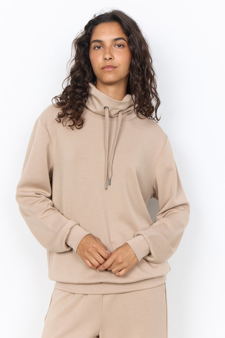 A woman with long, curly dark brown hair wearing a beige pullover hoodie with a drawstring and matching sweatpants, standing against a plain white studio background.