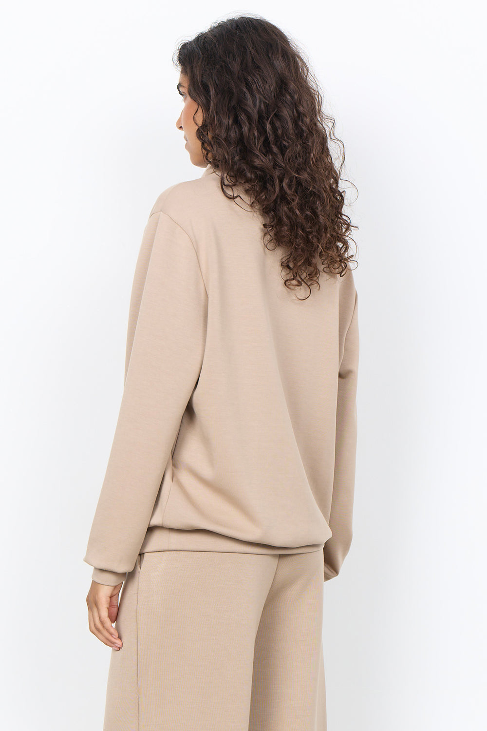 A woman with long, curly brown hair is shown from the back wearing a beige knit crewneck sweatshirt and matching beige wide-leg pants against a plain white studio background.