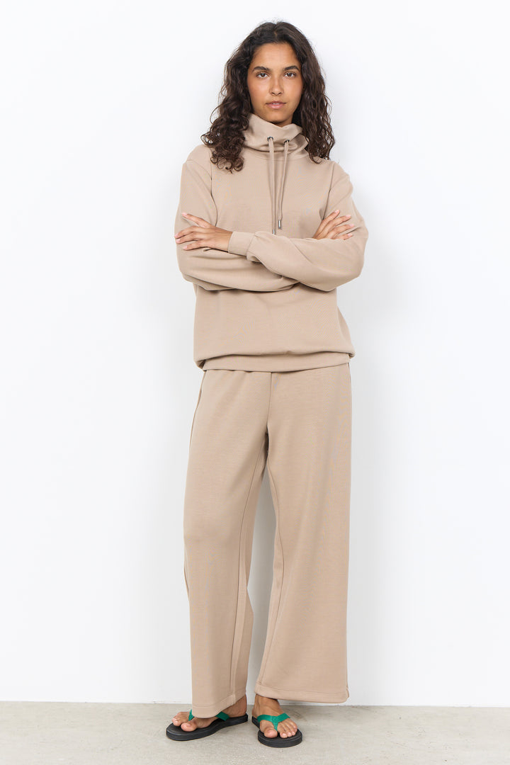 A woman modeling a matching beige lounge set consisting of a zip-up hoodie and wide-leg sweatpants, paired with green sandals, against a plain white wall.