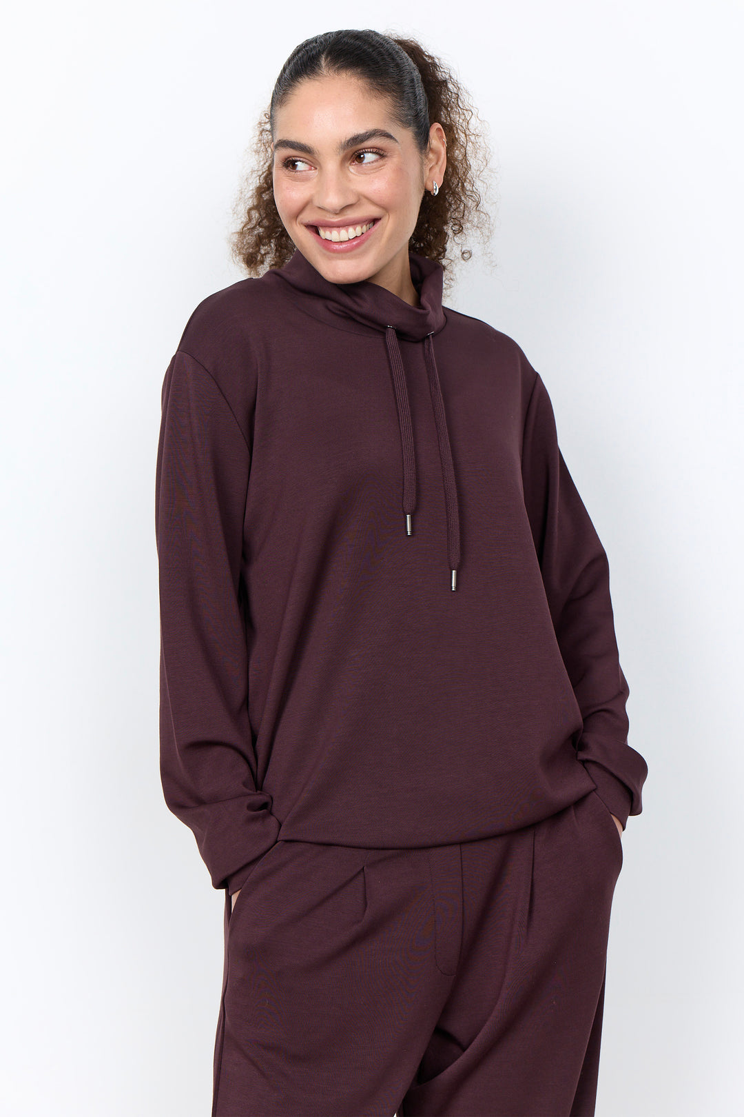 A woman wearing a matching dark plum-brown lounge set consisting of a high-neck sweatshirt with drawstrings and coordinating sweatpants, photographed against a clean white studio background.