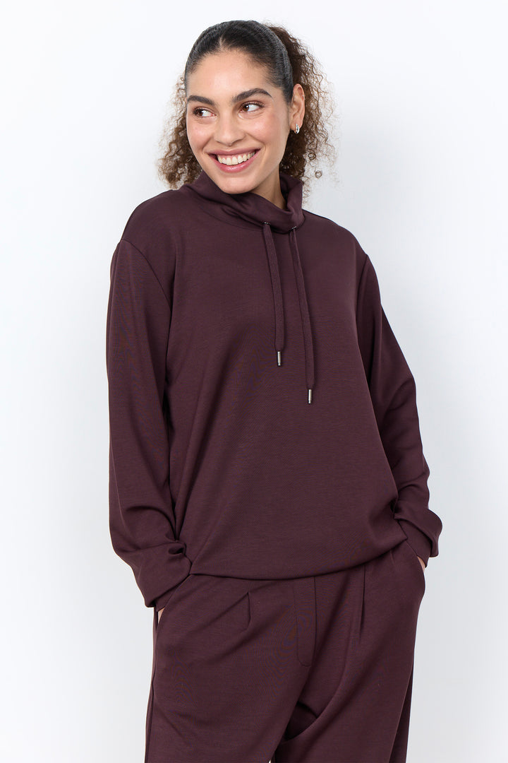 A woman wearing a matching dark plum-brown lounge set consisting of a high-neck sweatshirt with drawstrings and coordinating sweatpants, photographed against a clean white studio background.