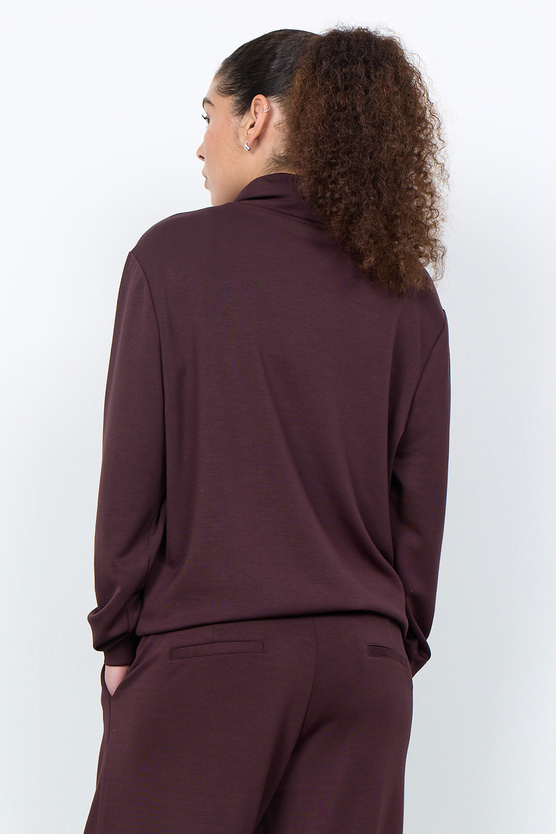 A woman with curly brown hair tied back, shown from behind wearing a matching dark plum-brown long-sleeve top and trousers set against a plain light gray studio background.