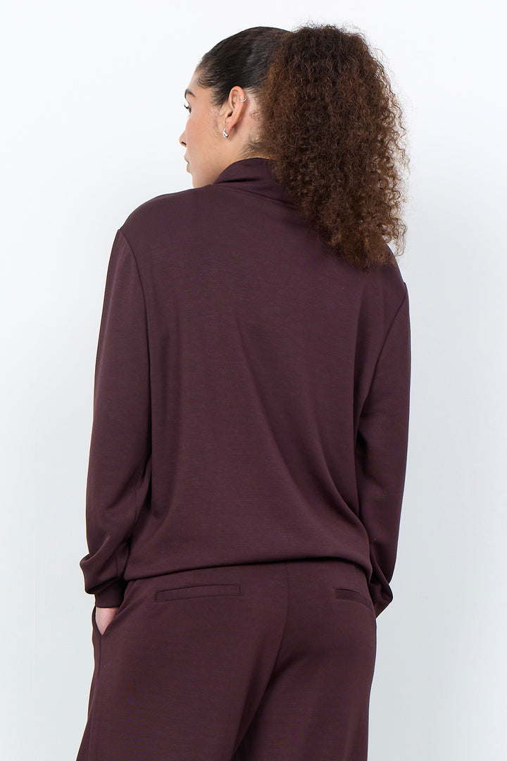 A woman with curly brown hair tied back, shown from behind wearing a matching dark plum-brown long-sleeve top and trousers set against a plain light gray studio background.