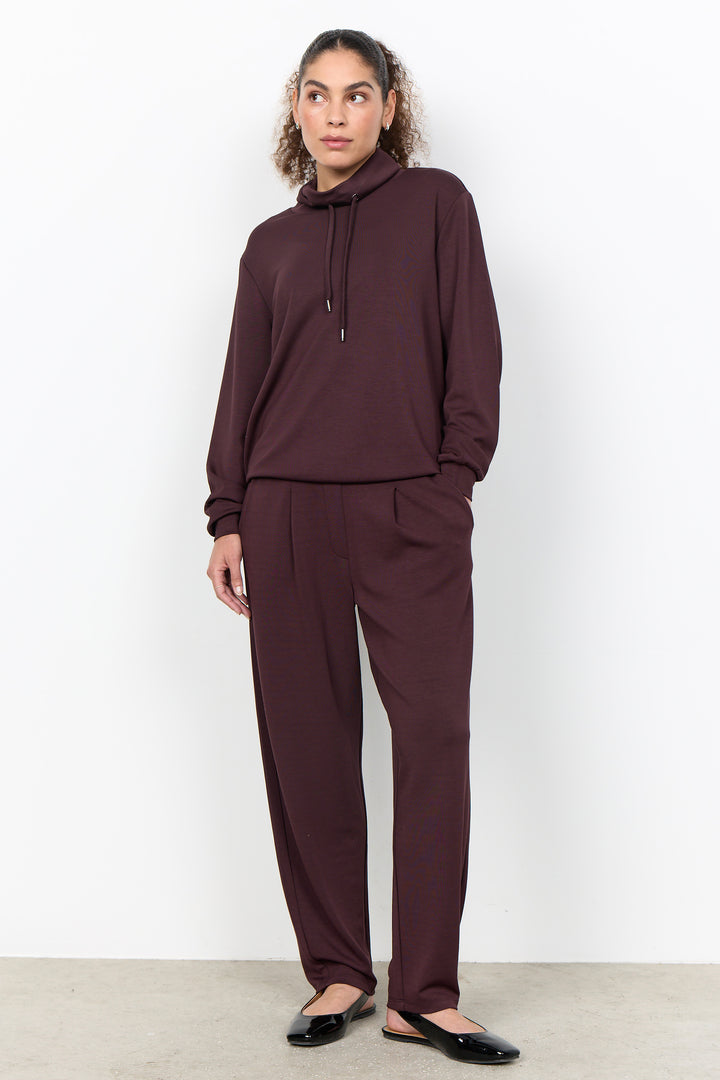 A woman modeling a matching two-piece loungewear set in a deep plum-burgundy color, featuring a drawstring pullover hoodie and wide-leg pants, paired with black flats against a clean white studio background.