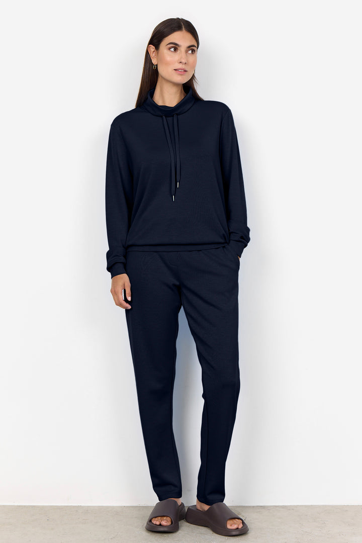 A woman modeling a matching dark navy lounge set consisting of a long-sleeve mock-neck hoodie with a drawstring and coordinating pants, paired with brown slide sandals against a plain white wall.