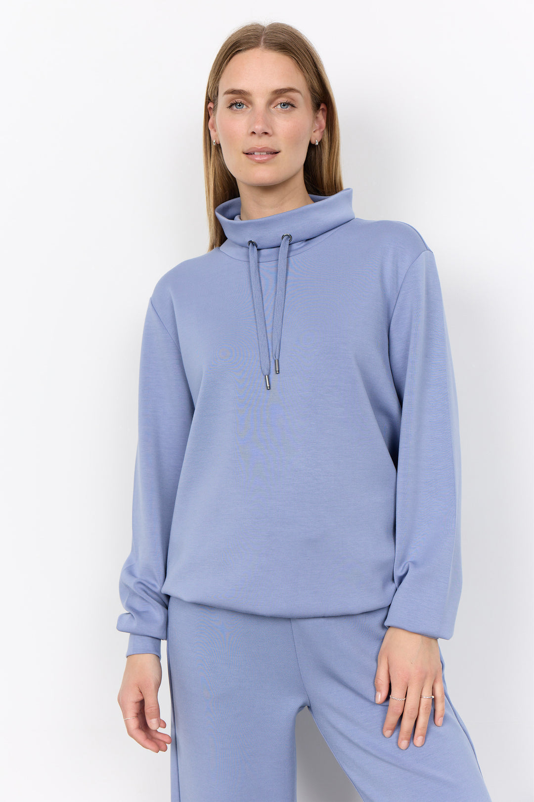 A woman modeling a light periwinkle blue sweatshirt and matching sweatpants set against a plain white studio background.