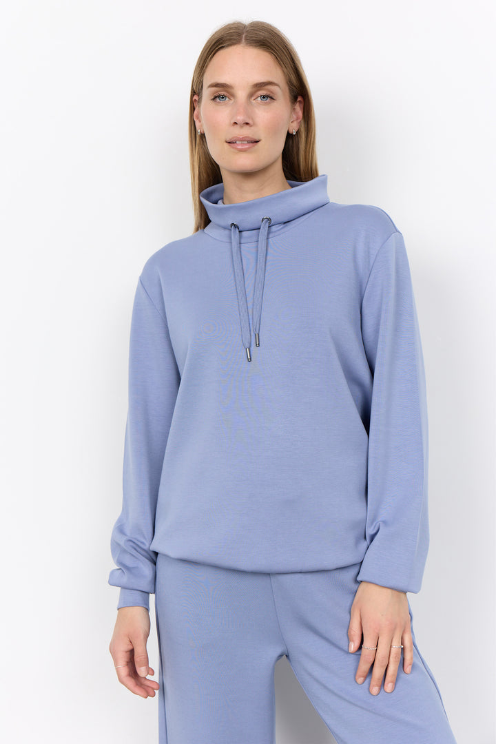 A woman modeling a light periwinkle blue sweatshirt and matching sweatpants set against a plain white studio background.