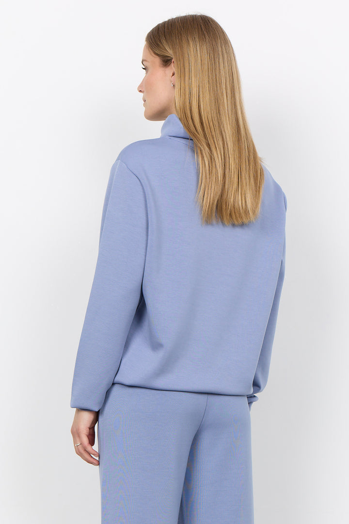 A woman with long blonde hair is shown from the back wearing a light periwinkle blue long-sleeve top and matching trousers against a plain white studio background.