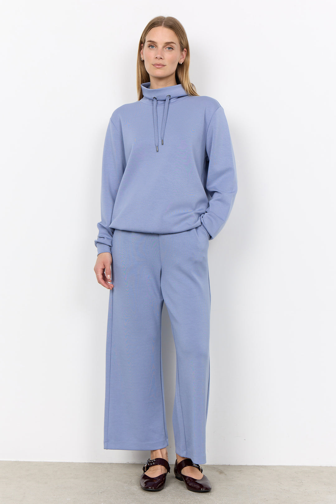 A woman modeling a matching dusty blue lounge set consisting of a long-sleeve mock-neck hoodie and wide-leg cropped pants, paired with dark brown flat shoes against a white wall.