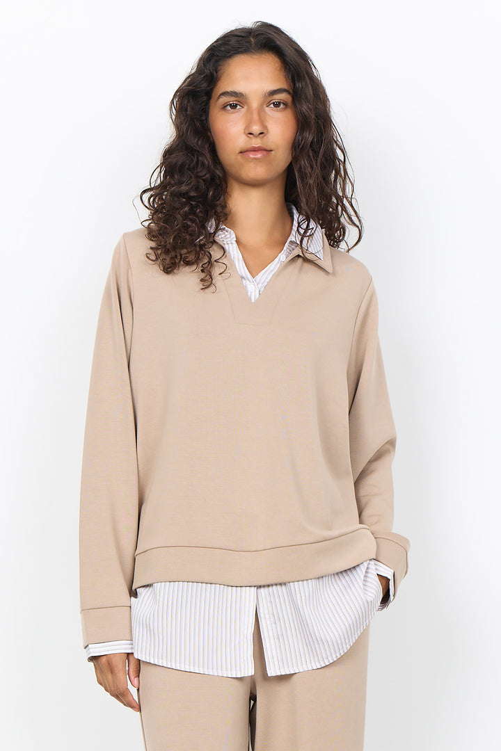A woman modeling a beige long-sleeve knit sweater with a split V-neck over a white collared shirt and striped shirt, paired with beige trousers, against a plain white studio background.