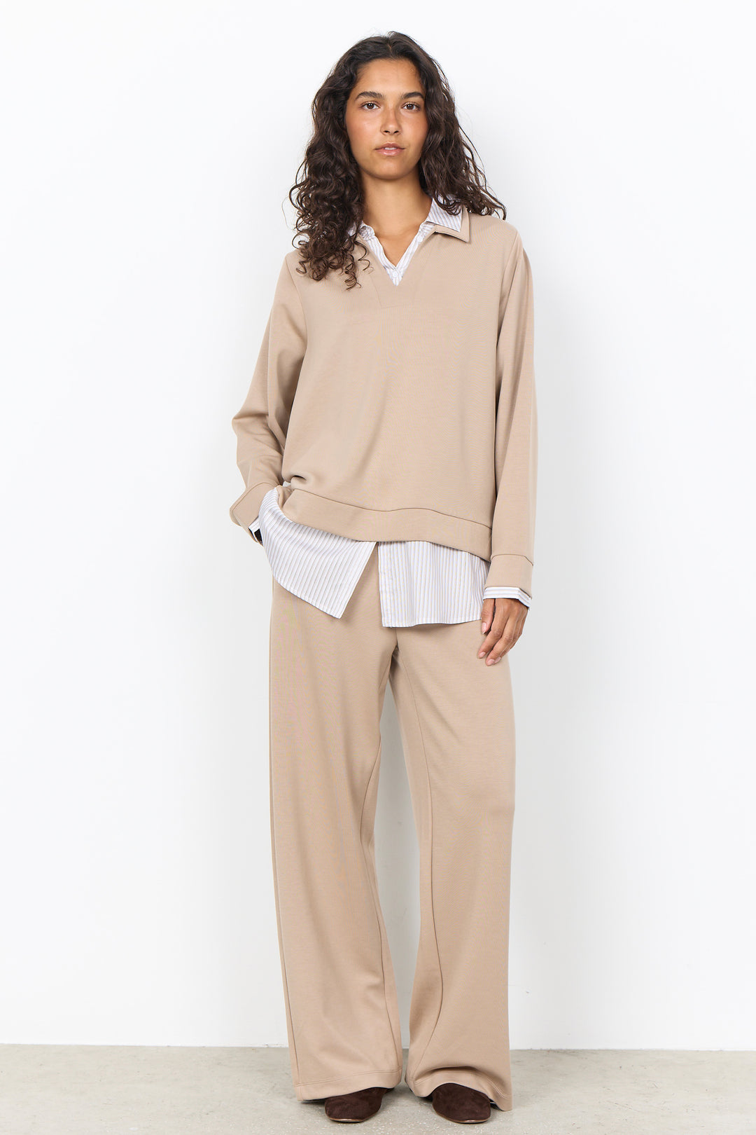 A model stands against a plain white studio wall wearing a beige knit long-sleeve top and matching wide-leg trousers, layered over a white collared shirt.