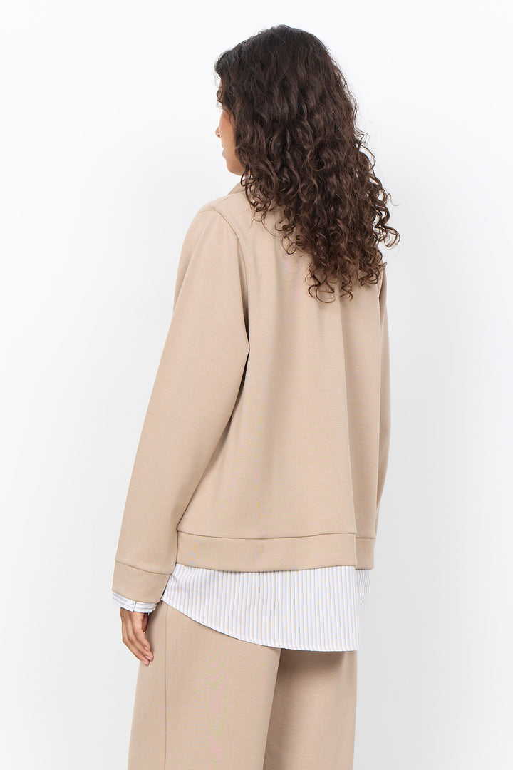 A woman with long, curly brown hair is shown from the back wearing a beige crewneck sweatshirt, matching beige trousers, and a white striped shirt underneath, set against a plain white studio background.