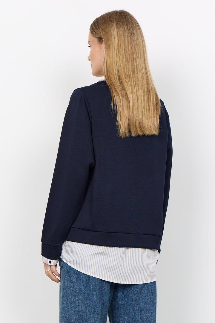 A woman with long blonde hair is shown from the back wearing a dark navy blue long-sleeve top over a white striped shirt and blue jeans, against a plain white background.