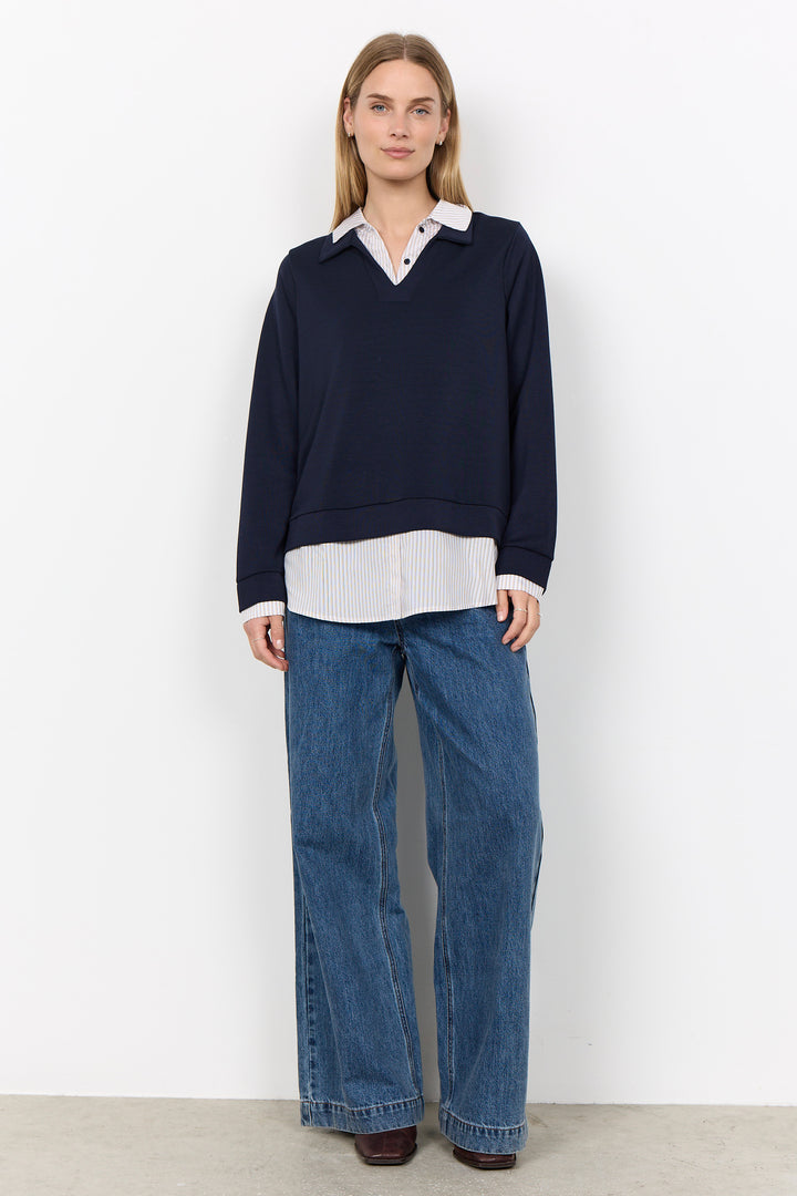 A woman stands against a plain white wall wearing a dark navy long-sleeve knit top layered over a white collared shirt, paired with wide-leg blue jeans and dark brown shoes.