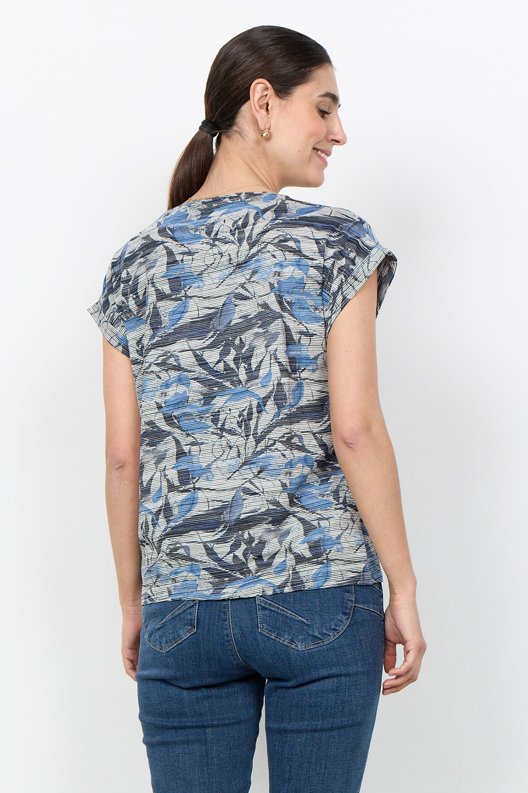 A woman shown from the back wearing a short-sleeve top with a blue, gray, and white abstract pattern, paired with dark blue jeans, against a plain white background.