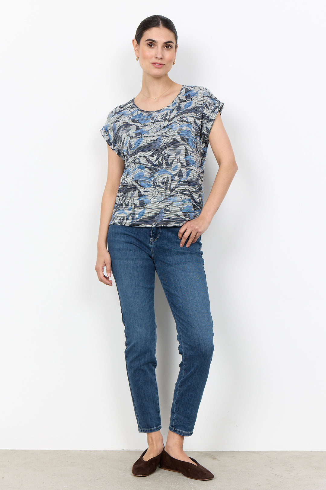 A woman modeling a short-sleeve t-shirt with a blue, gray, and white abstract pattern, paired with blue jeans and dark brown flat shoes, standing against a plain white wall.