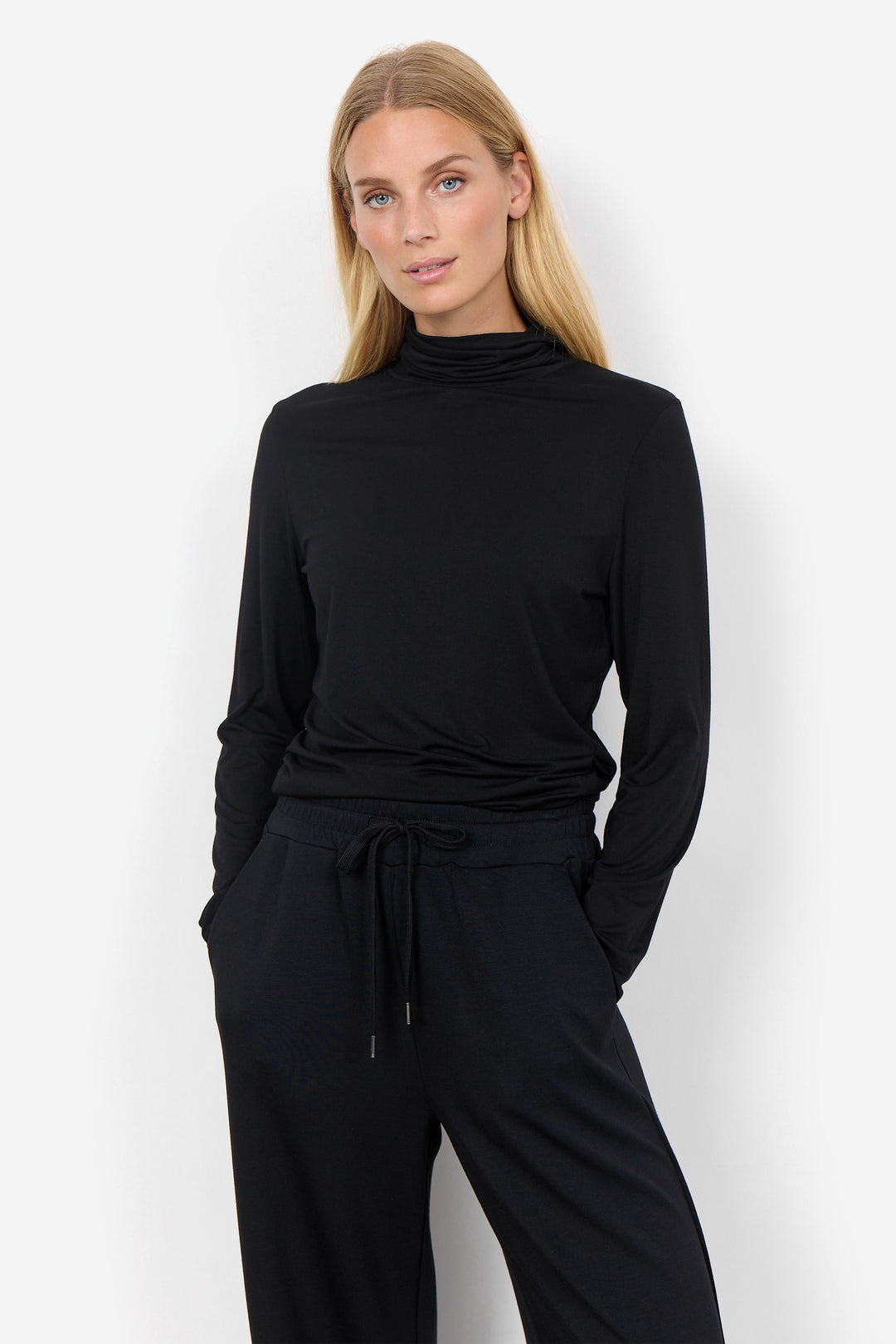 A blonde woman modeling a black long-sleeve mock neck top and matching black drawstring pants against a plain white studio background.