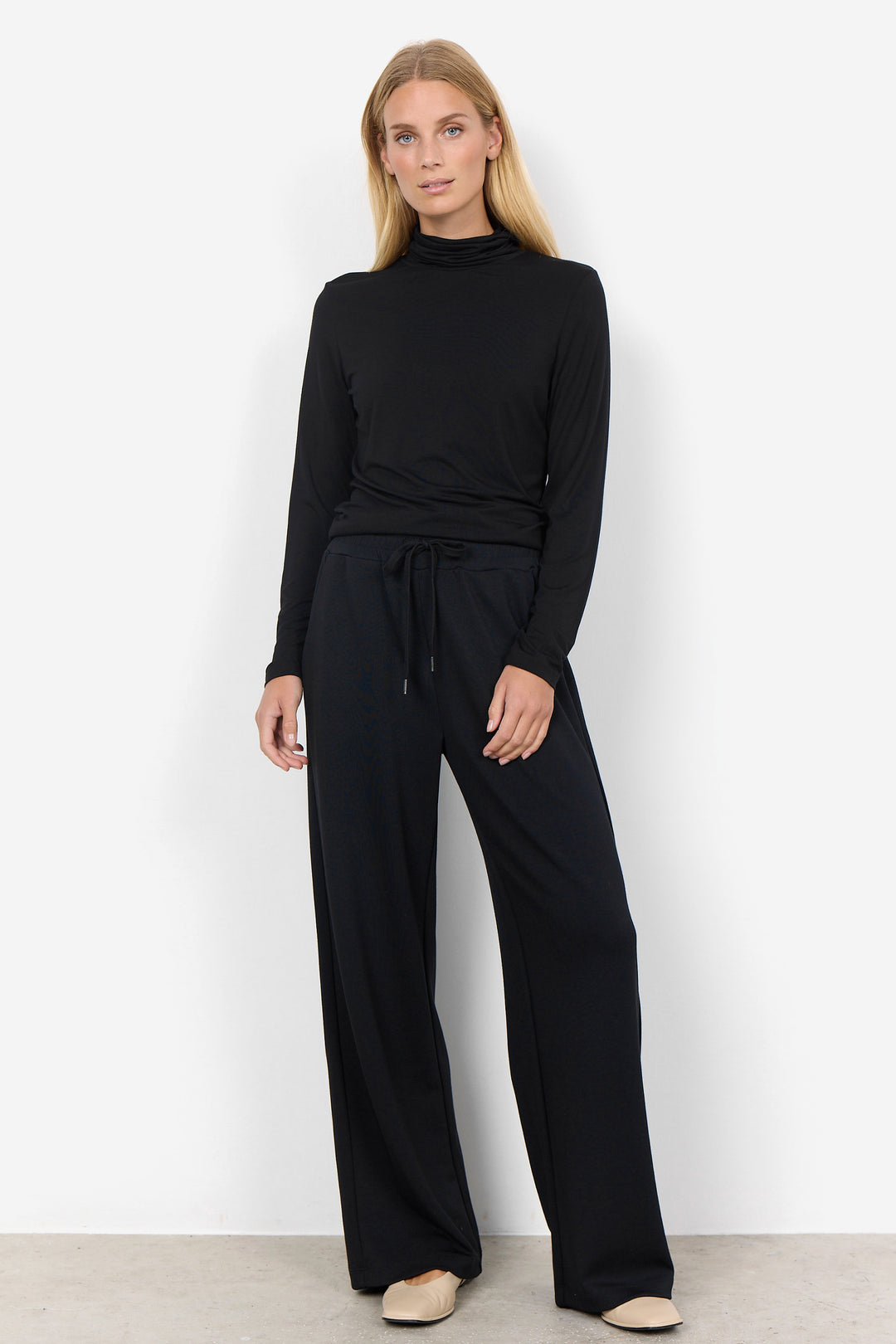 A woman modeling a black long-sleeve mock-neck top and matching black wide-leg trousers with a drawstring waist, standing against a plain white studio wall.