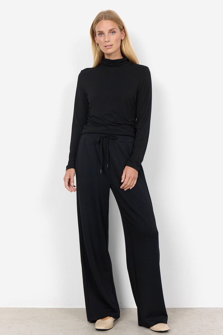 A woman modeling a black long-sleeve mock-neck top and matching black wide-leg trousers with a drawstring waist, standing against a plain white studio wall.