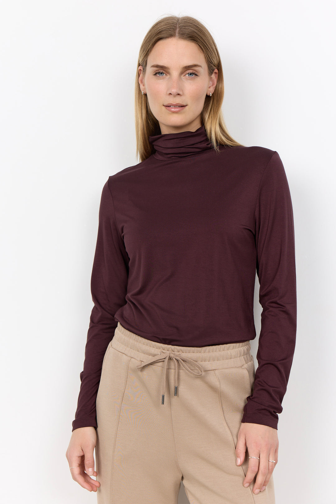 A woman modeling a dark burgundy long-sleeve mock neck top paired with beige drawstring pants against a plain white studio background.