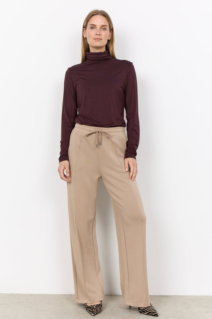 A woman modeling a dark burgundy long-sleeve turtleneck top and beige wide-leg drawstring trousers against a plain white studio background.