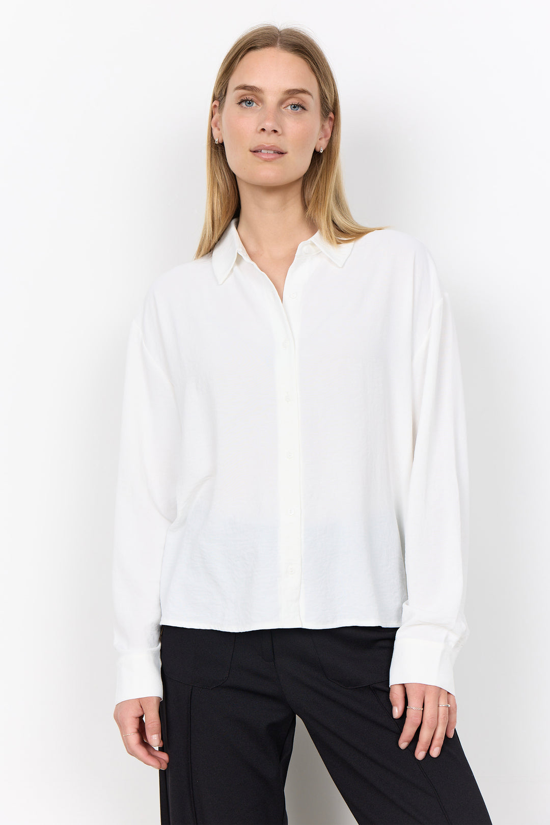 A woman modeling a white long-sleeve button-up blouse with a relaxed fit, paired with black trousers, against a plain white studio background.