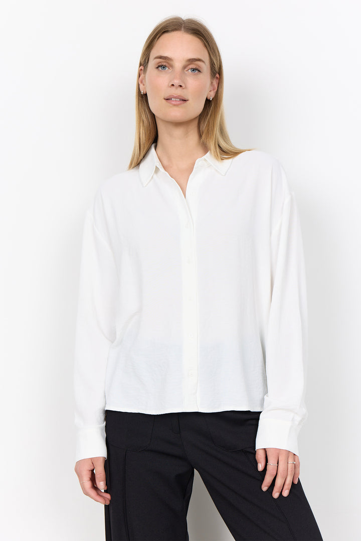 A woman modeling a white long-sleeve button-up blouse with a relaxed fit, paired with black trousers, against a plain white studio background.