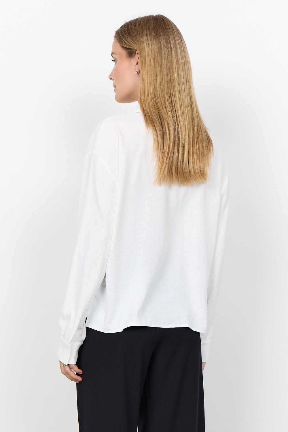 A woman with long blonde hair is shown from the back wearing a white long-sleeve blouse and black trousers against a plain white background.