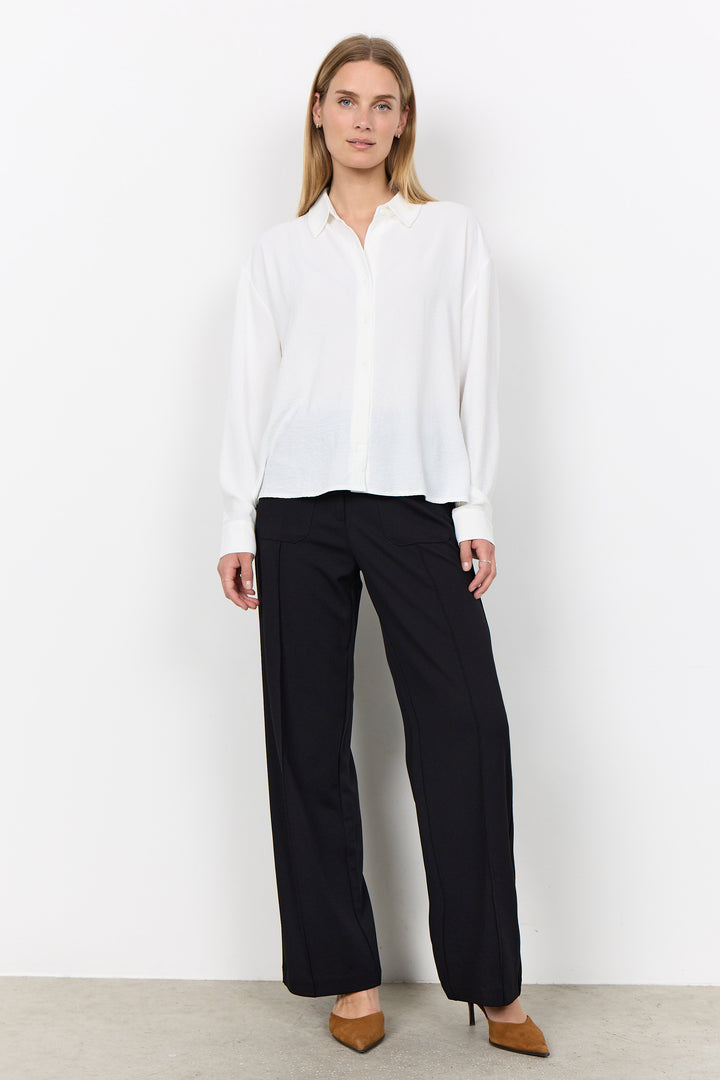 A woman modeling a white long-sleeve button-up blouse and black wide-leg trousers, standing against a plain white studio wall.