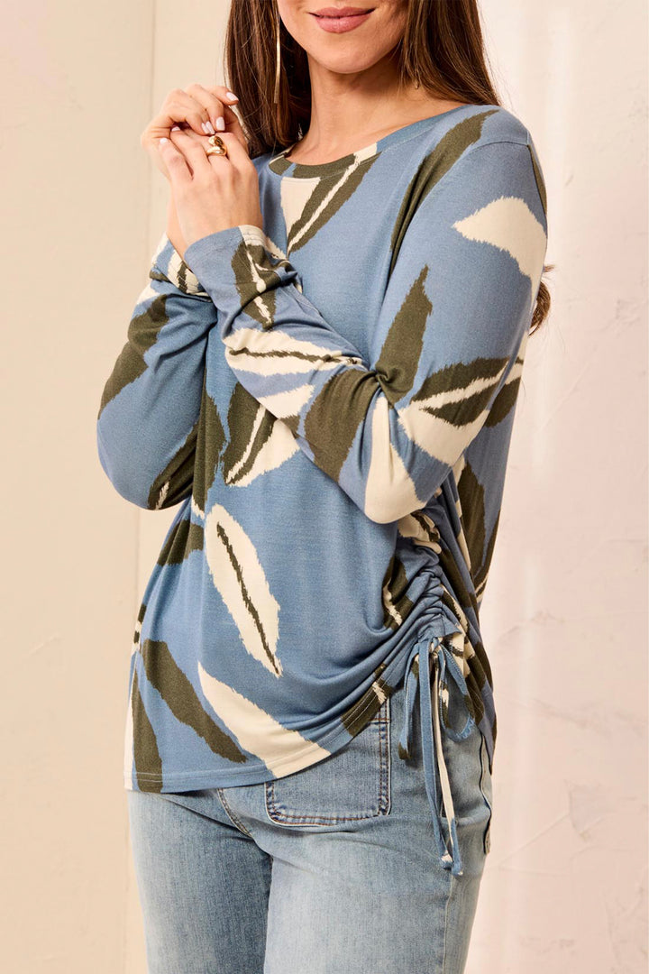 Woman wearing a blue long-sleeve top with leaf pattern and tied front, standing against a beige background.